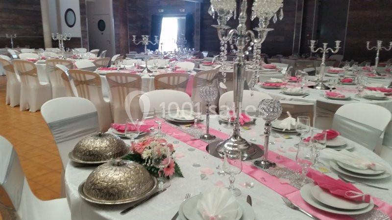 Salle de réception décorée avec des tables dressées, nappes blanches, chemins de table roses et chandeliers argentés.
