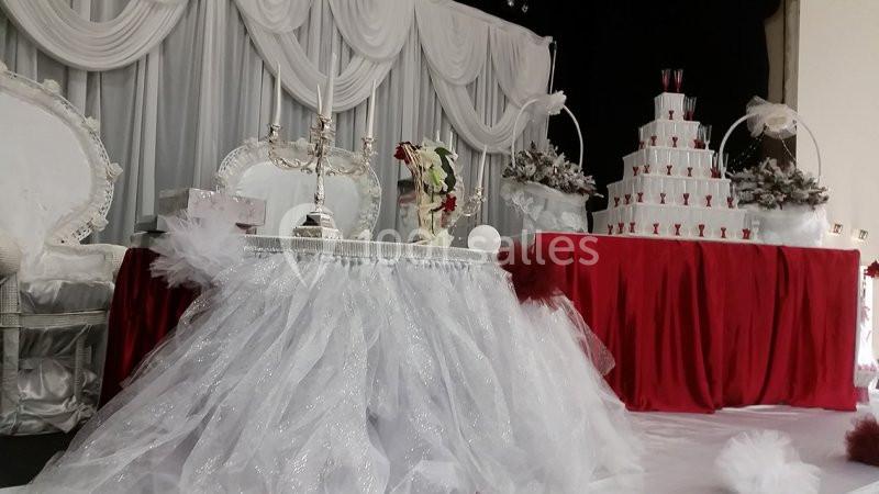Décoration de mariage avec table ornée de tulle blanc, chandelier, fleurs et fontaine de coupes sur fond rouge.