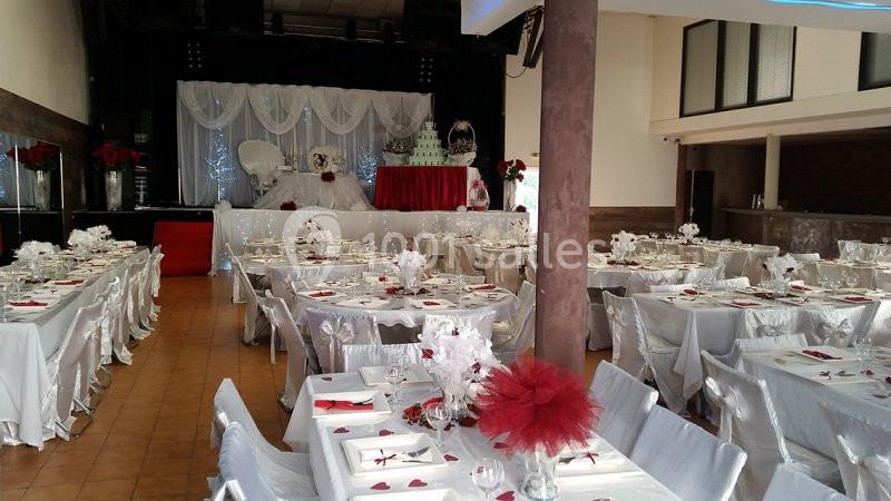 Salle de réception décorée pour un mariage, avec tables dressées en blanc et rouge et une scène ornée d'un gâteau.