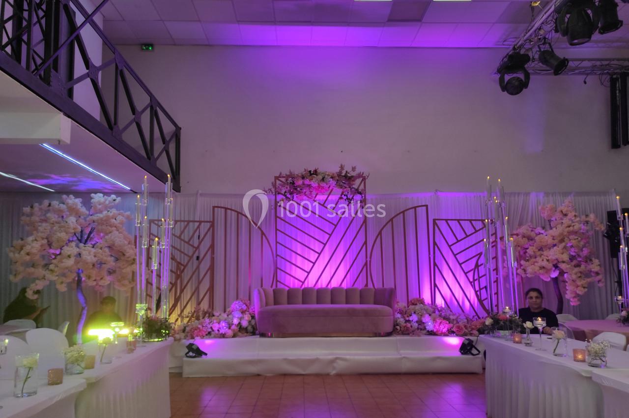 Décor de salle avec canapé rose, éclairage violet, fleurs et bougies, entouré de tables dressées pour un événement.