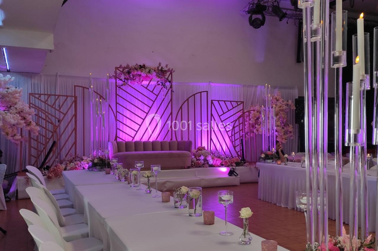Salle de réception décorée avec des tons roses et violets, tables dressées et bougies hautes illuminées.