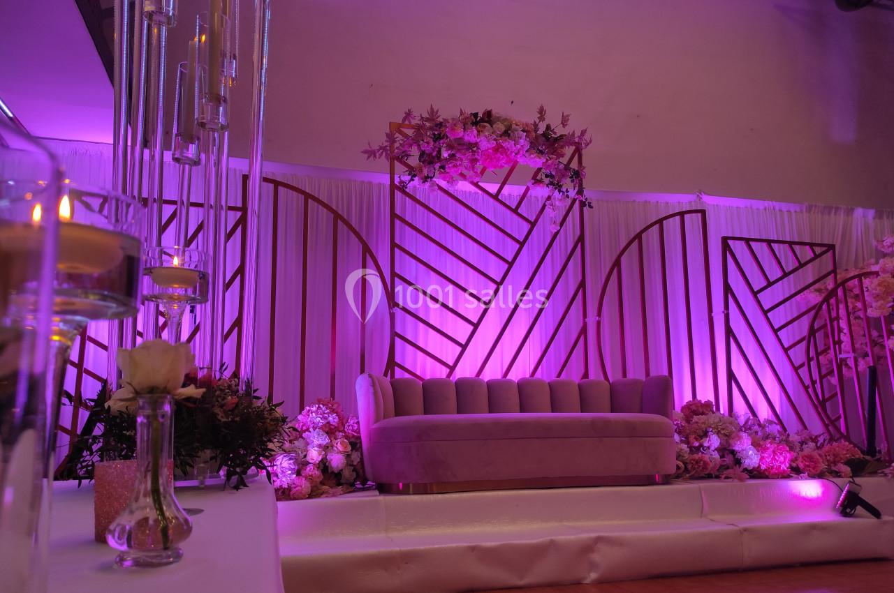 Décor de scène avec canapé rose, fleurs et éclairage violet, entouré de structures géométriques et bougies.