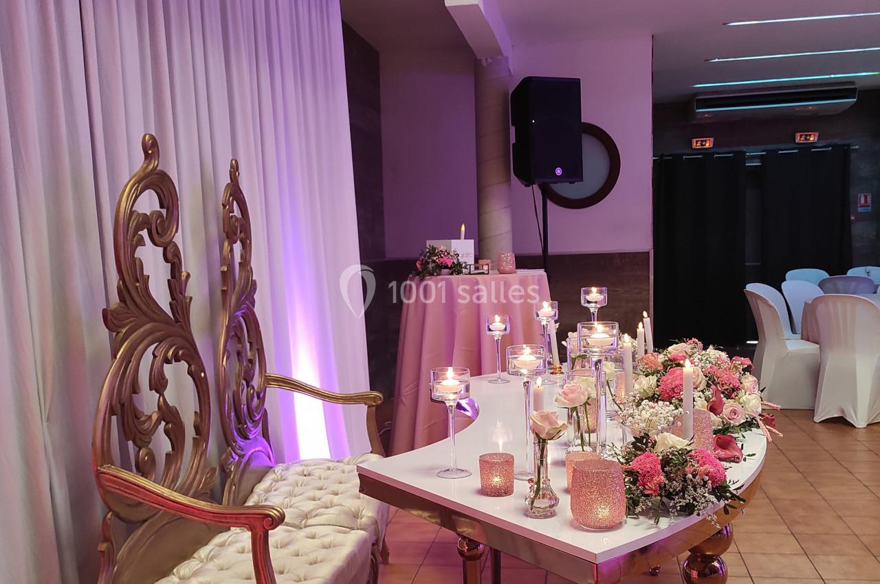 Chaises dorées ornées et table décorée de bougies et fleurs dans une salle de réception aux tons roses et blancs.