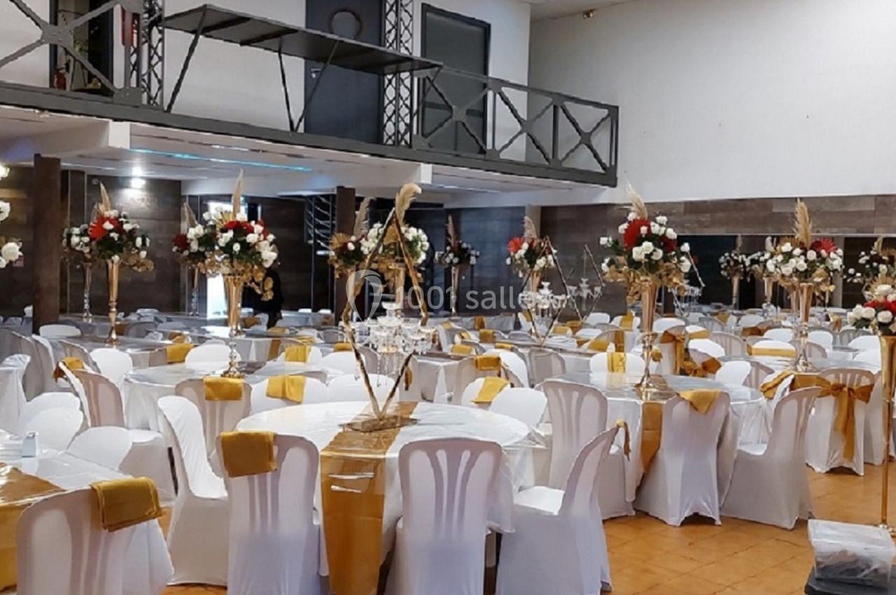 Salle de réception décorée avec des tables dressées, nappes blanches, chemins de table dorés et arrangements floraux.