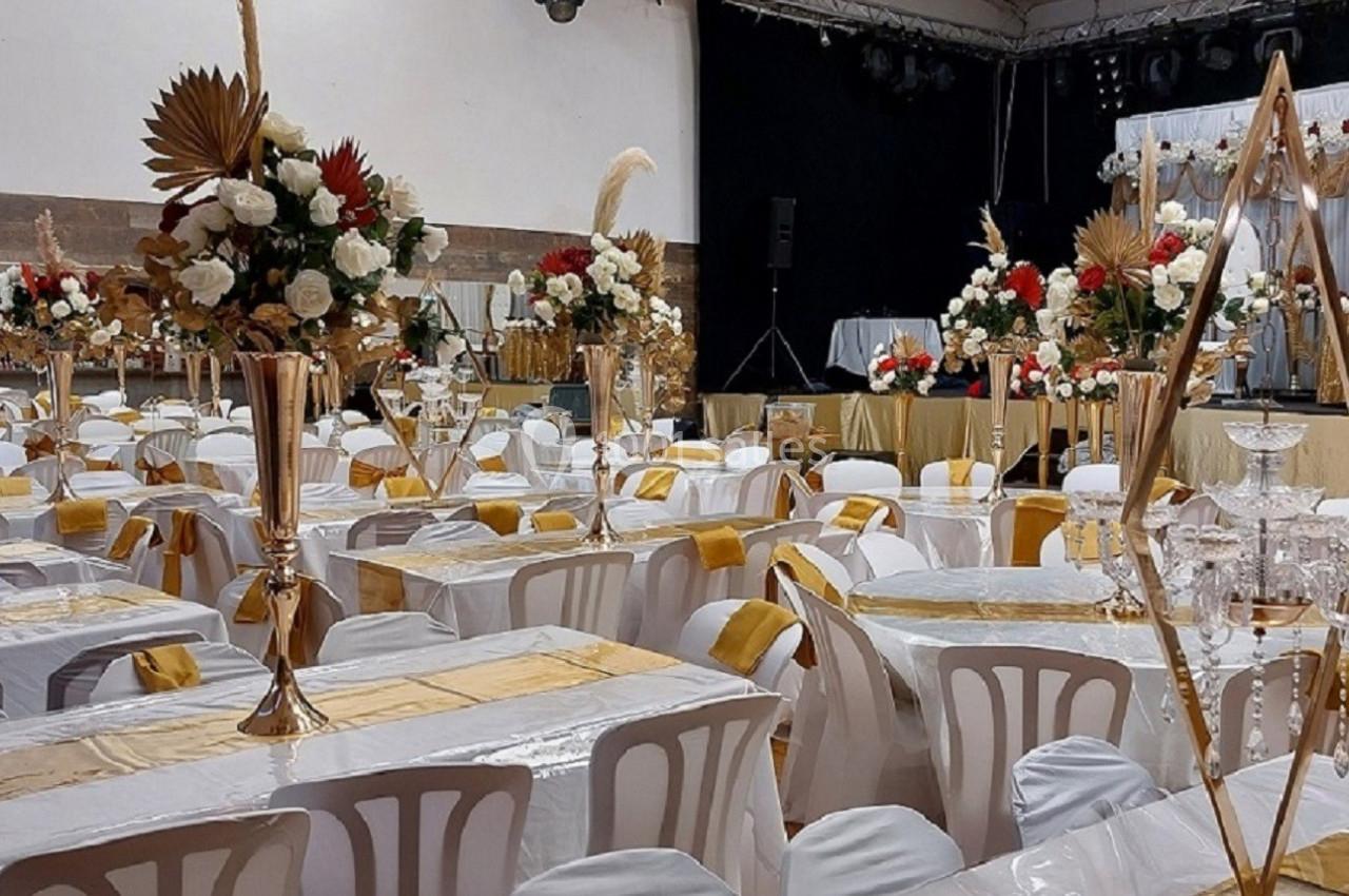 Salle de réception décorée avec des tables alignées, nappes blanches et dorées, et centres de table floraux élégants.