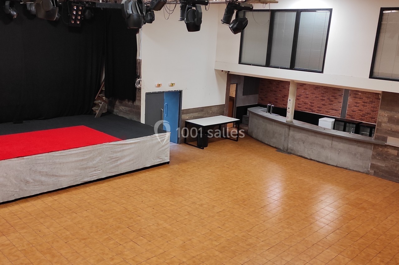 Salle polyvalente avec scène surélevée, rideaux noirs, projecteurs, comptoir en angle et sol en carrelage.