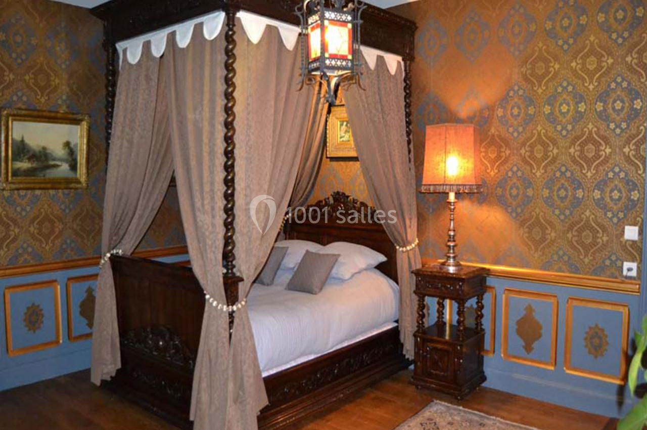 Chambre Louis XIII Lit à baldaquin en bois sculpté avec rideaux, dans une chambre décorée de motifs classiques et éclairée par une lampe.