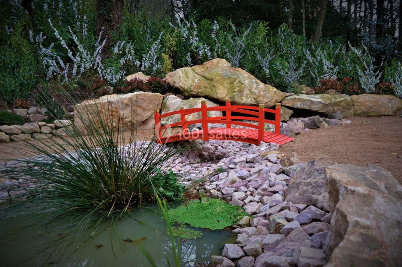 Partie du jardin chinois Petit pont rouge en bois traversant un ruisseau entouré de pierres, de végétation et d'un étang avec des plantes aquatiques.