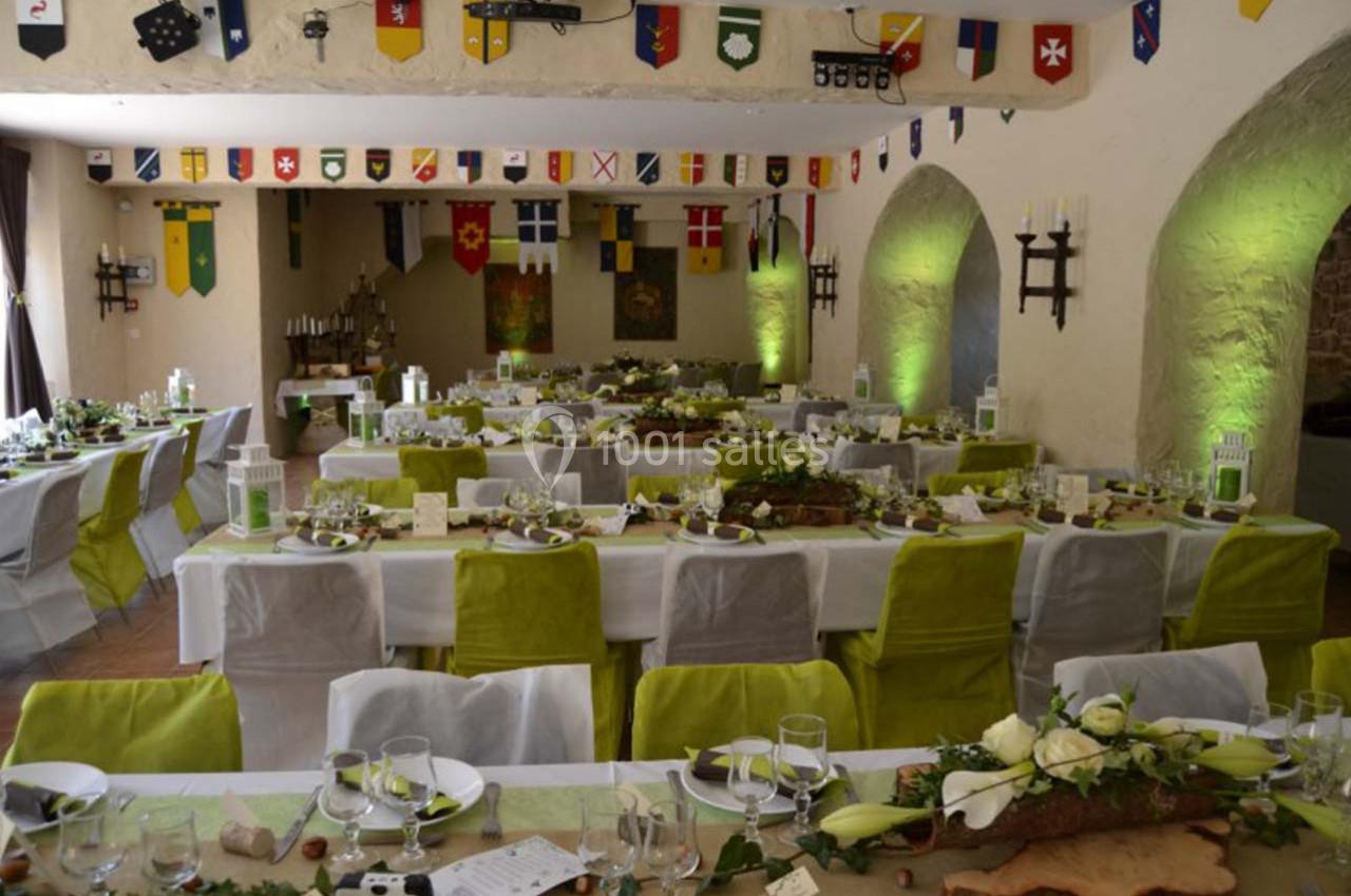 Salle de mariage 110 personnes autour de tables Salle décorée pour un banquet, avec des tables dressées, des chaises vertes et des bannières médiévales suspendues.