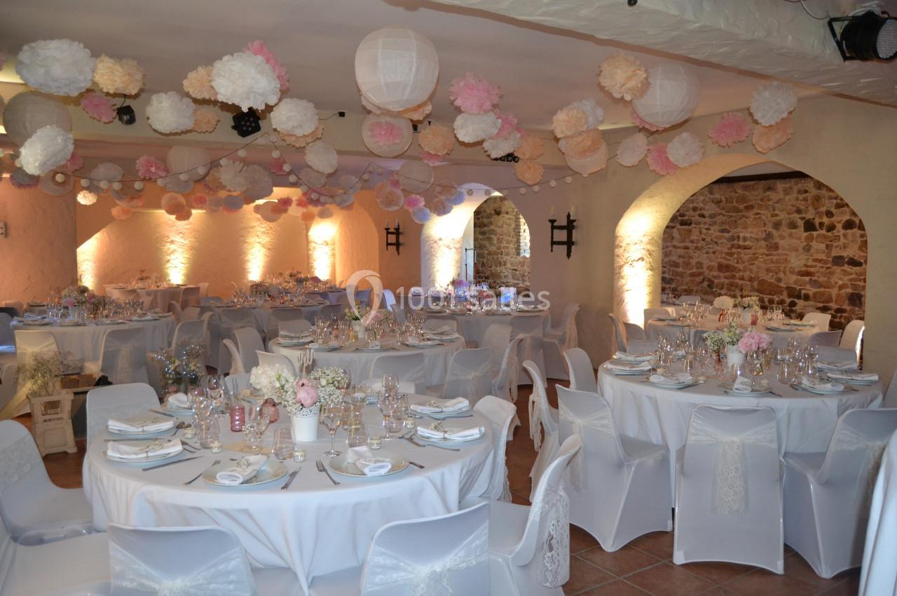 Salle de mariage 110 personnes autour de tables Salle décorée pour un mariage avec tables rondes, nappes blanches, vaisselle élégante et guirlandes de lanternes et pompons…