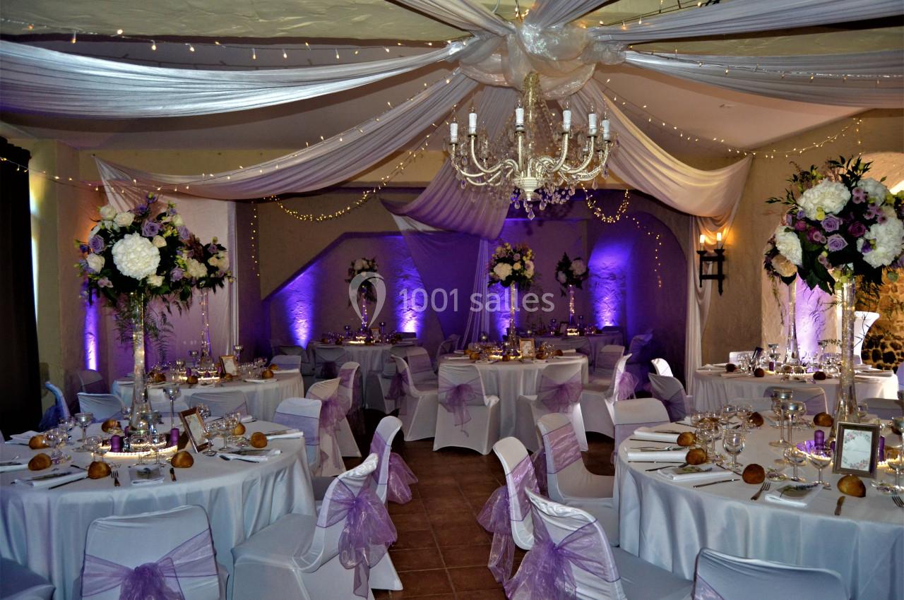 Salle de mariage 110 personnes autour de tables Salle de réception décorée avec des nappes blanches, des rubans violets, des fleurs et un éclairage tamisé.