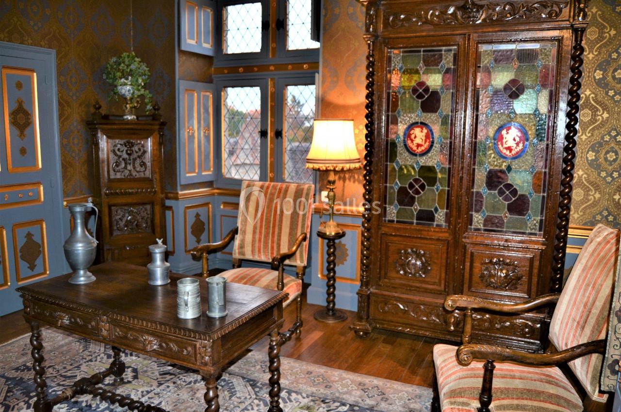 Salon chambre Louix XIII Salle de style ancien avec mobilier en bois sculpté, vitrail coloré, tapis et chaises rembourrées à rayures.