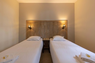 Deux lits simples avec draps blancs, séparés par une table de chevet en bois, éclairés par deux appliques murales.