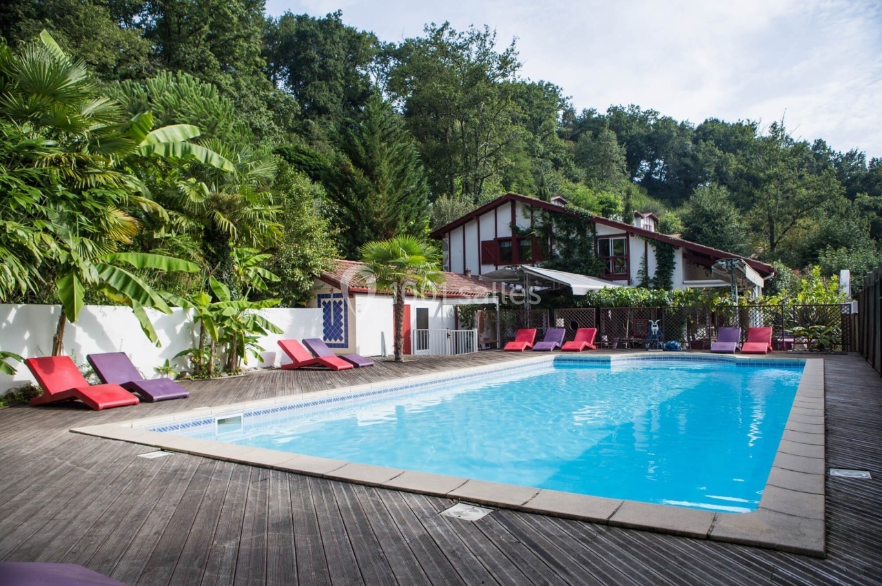 Piscine extérieure entourée de transats colorés, avec une maison traditionnelle et des arbres en arrière-plan.