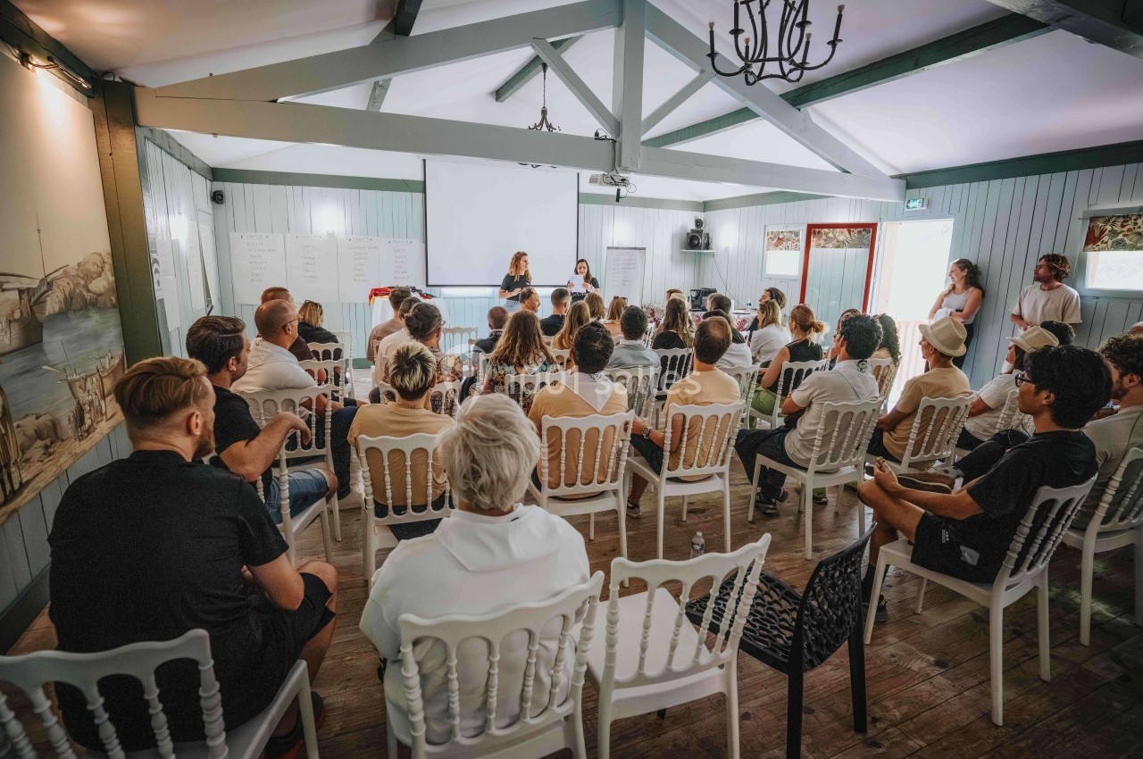 Groupe de personnes assises dans une salle, écoutant une présentation devant un écran et des tableaux blancs.