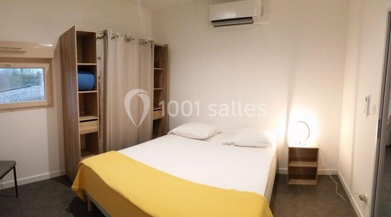 Chambre avec lit double, couverture jaune, placard ouvert, petite fenêtre et éclairage d'appoint.