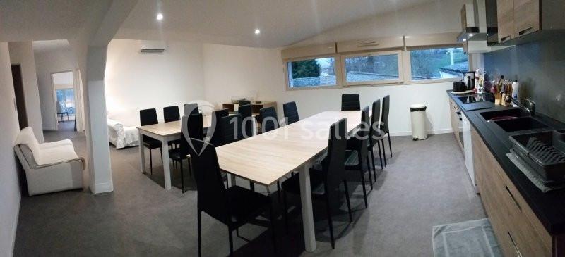 Salle lumineuse avec une grande table en bois, des chaises noires, une cuisine équipée et des fenêtres donnant sur l…