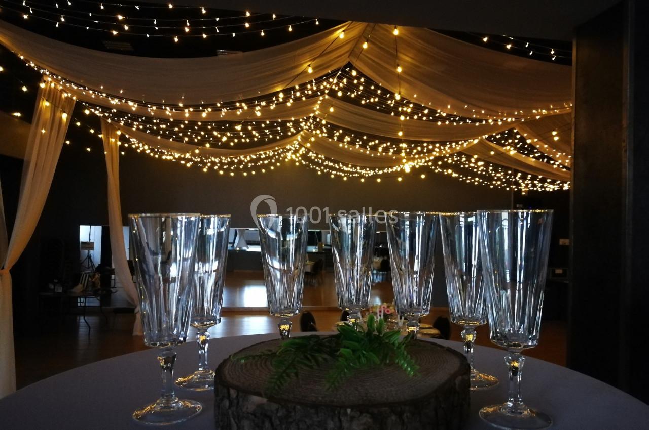 Des verres en cristal posés sur une table décorée, avec un plafond illuminé de guirlandes lumineuses et de drapés.