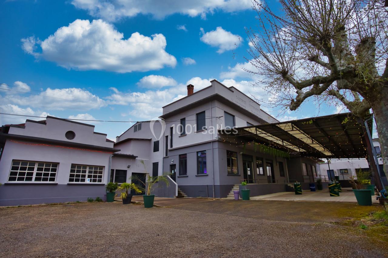Bâtiment industriel rénové avec façade grise, grande verrière et arbres sans feuilles sous un ciel partiellement nuageux.