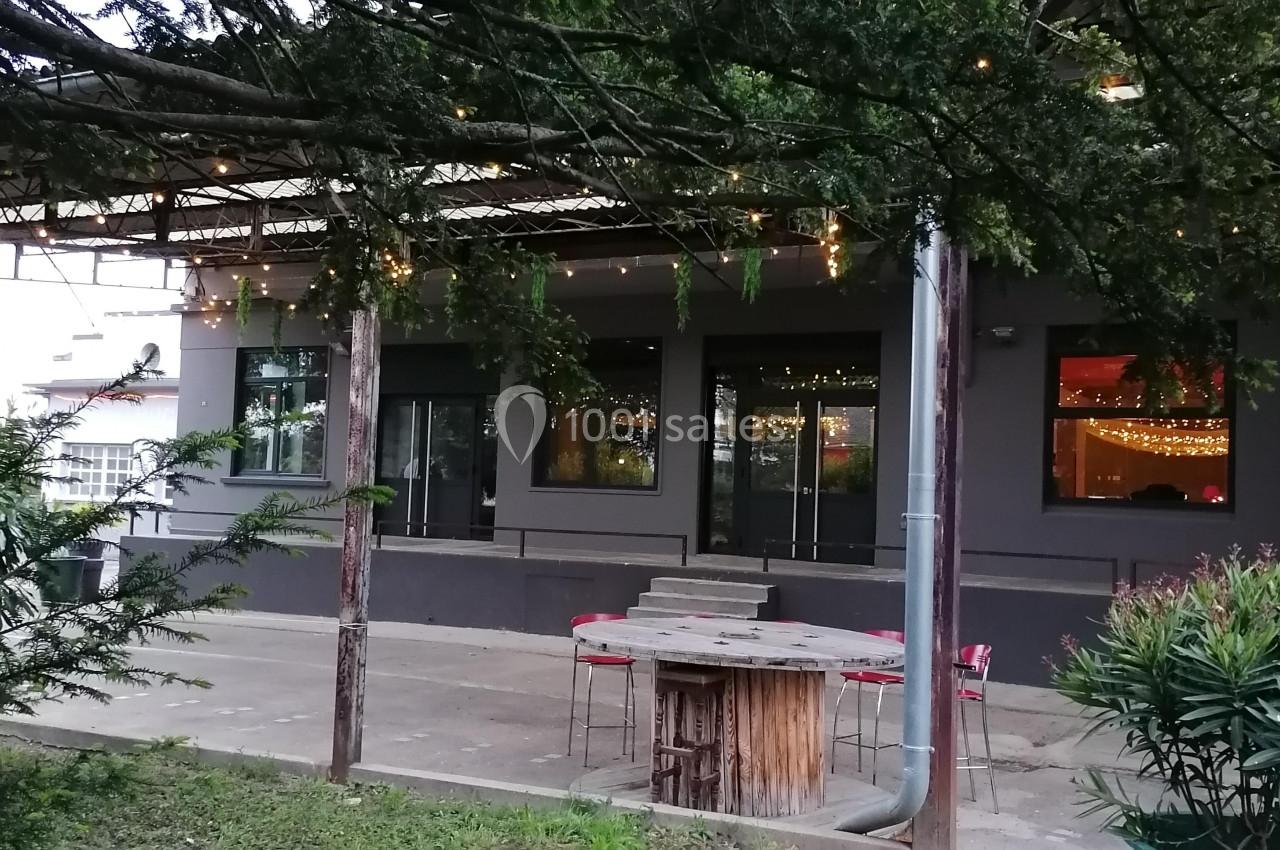 Terrasse extérieure avec tables, chaises et guirlandes lumineuses sous un auvent, entourée de végétation.