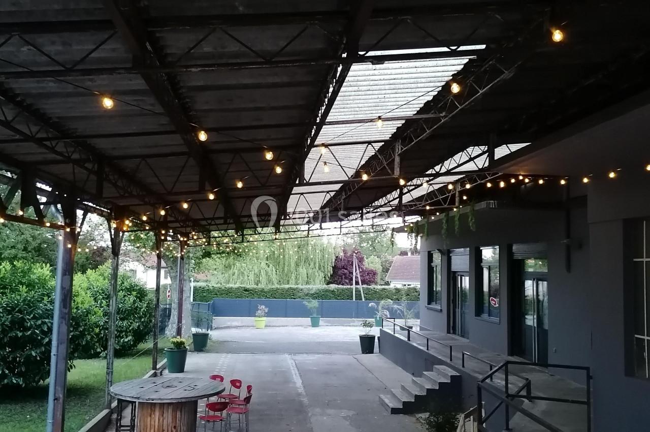 Terrasse couverte avec guirlandes lumineuses, table ronde en bois, chaises rouges et vue sur un jardin arboré.