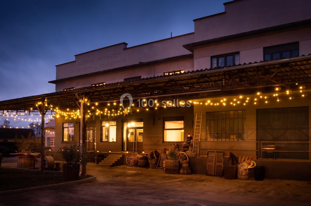 Façade d'un bâtiment industriel éclairé par des guirlandes lumineuses au crépuscule, avec des caisses et tonneaux devant.