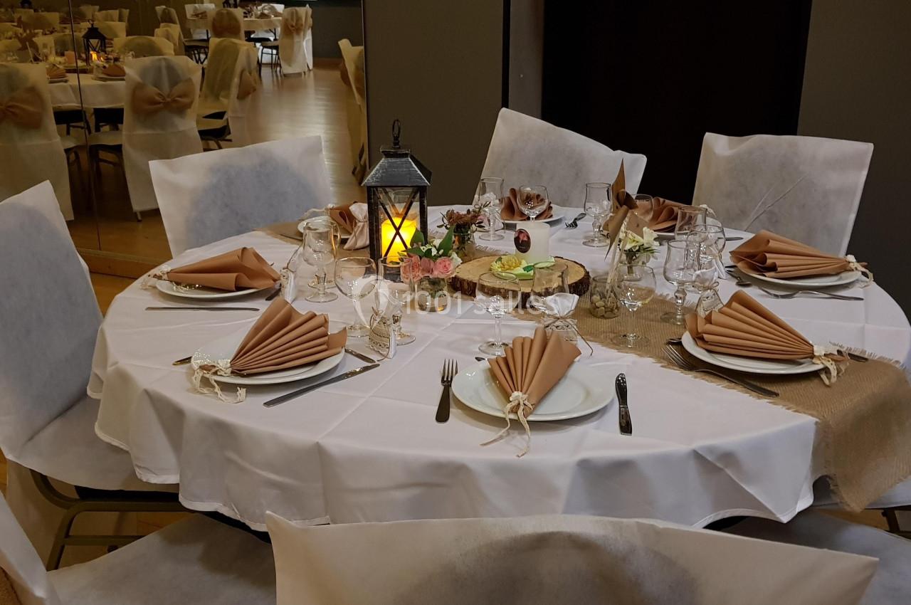 Table ronde élégamment dressée avec nappes blanches, serviettes pliées beige, lanternes et décorations rustiques.