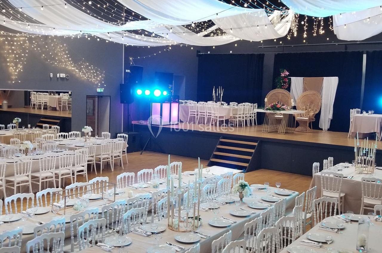 Salle de réception décorée pour un événement, avec tables dressées, chaises blanches et éclairage festif suspendu.