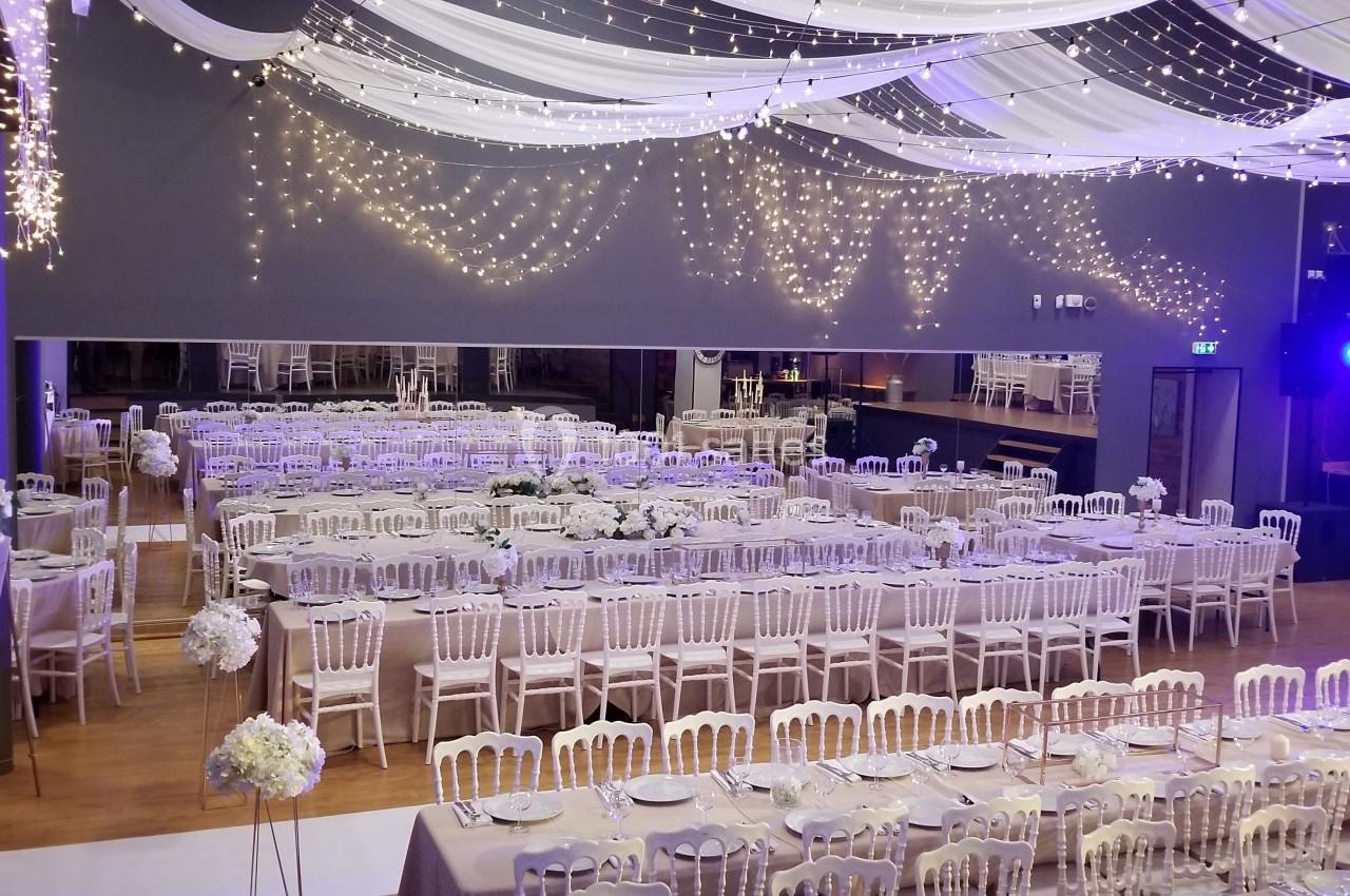 Salle de réception décorée avec des guirlandes lumineuses, tables dressées et chaises blanches alignées.