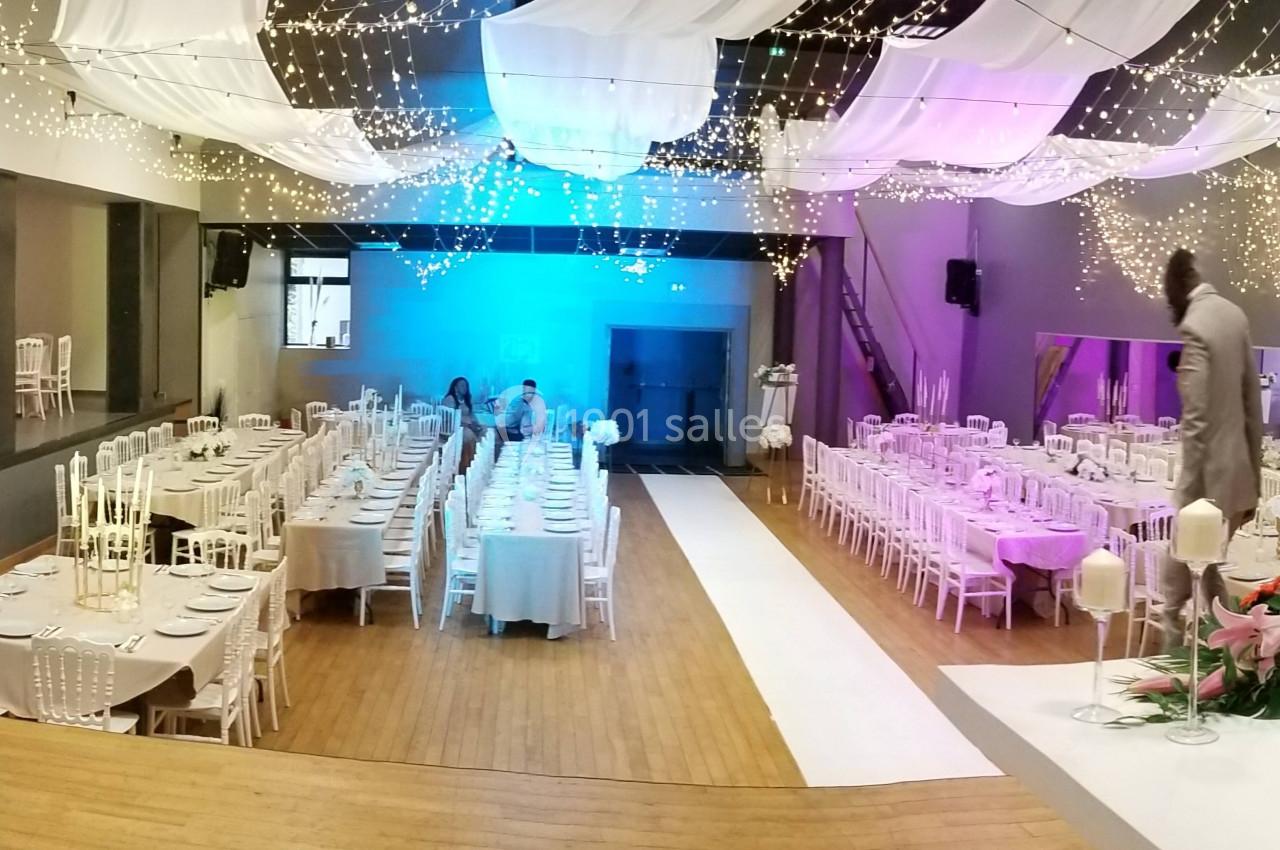 Salle de réception décorée pour un mariage, avec tables alignées, guirlandes lumineuses et une piste centrale blanche.