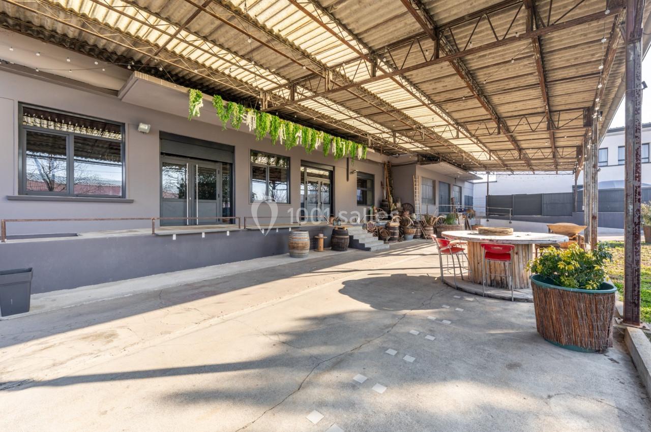 Terrasse couverte avec tables, chaises, plantes en pot et décorations suspendues, devant un bâtiment gris.