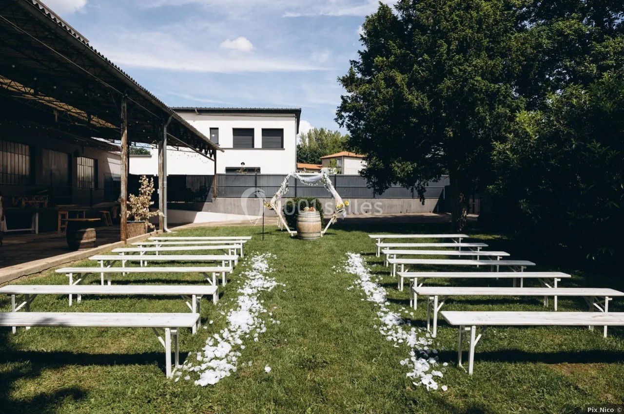 Allée centrale décorée de pétales blancs, entourée de bancs en bois, menant à une arche pour une cérémonie en extérieur.