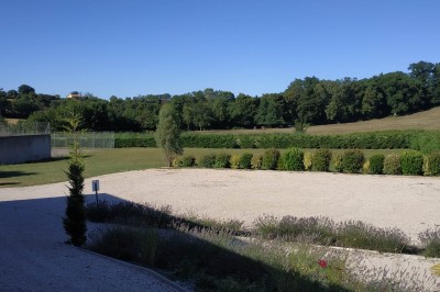 Vue d'un jardin avec allée gravillonnée, haies taillées, pelouse et arbres en arrière-plan sous un ciel dégagé.