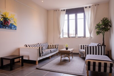 Salon lumineux avec canapé et fauteuils rayés, table basse en rotin, plante verte et grande fenêtre avec rideaux blancs.