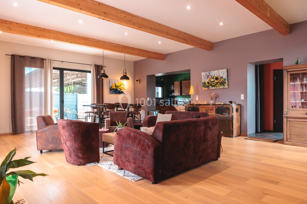 Salon spacieux avec parquet clair, canapés en velours marron, poutres apparentes et lumière naturelle abondante.