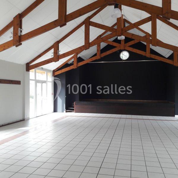 Salle vide avec plafond en poutres apparentes, sol carrelé blanc et estrade en bois devant un mur noir.