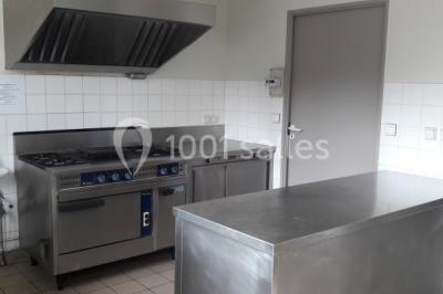 Cuisine professionnelle avec plan de travail en inox, cuisinière équipée et hotte, éclairée par une fenêtre.