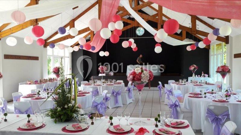 Salle de réception décorée avec des tables rondes, nappes blanches, rubans violets et lanternes suspendues roses et blanches.