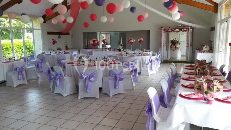 Salle décorée pour un événement avec des tables rondes et rectangulaires ornées de nappes blanches et rubans violets.