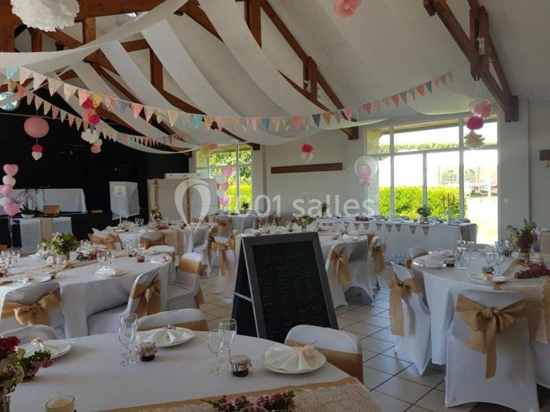 Salle de réception décorée pour un événement, avec tables dressées, guirlandes colorées et lumière naturelle.