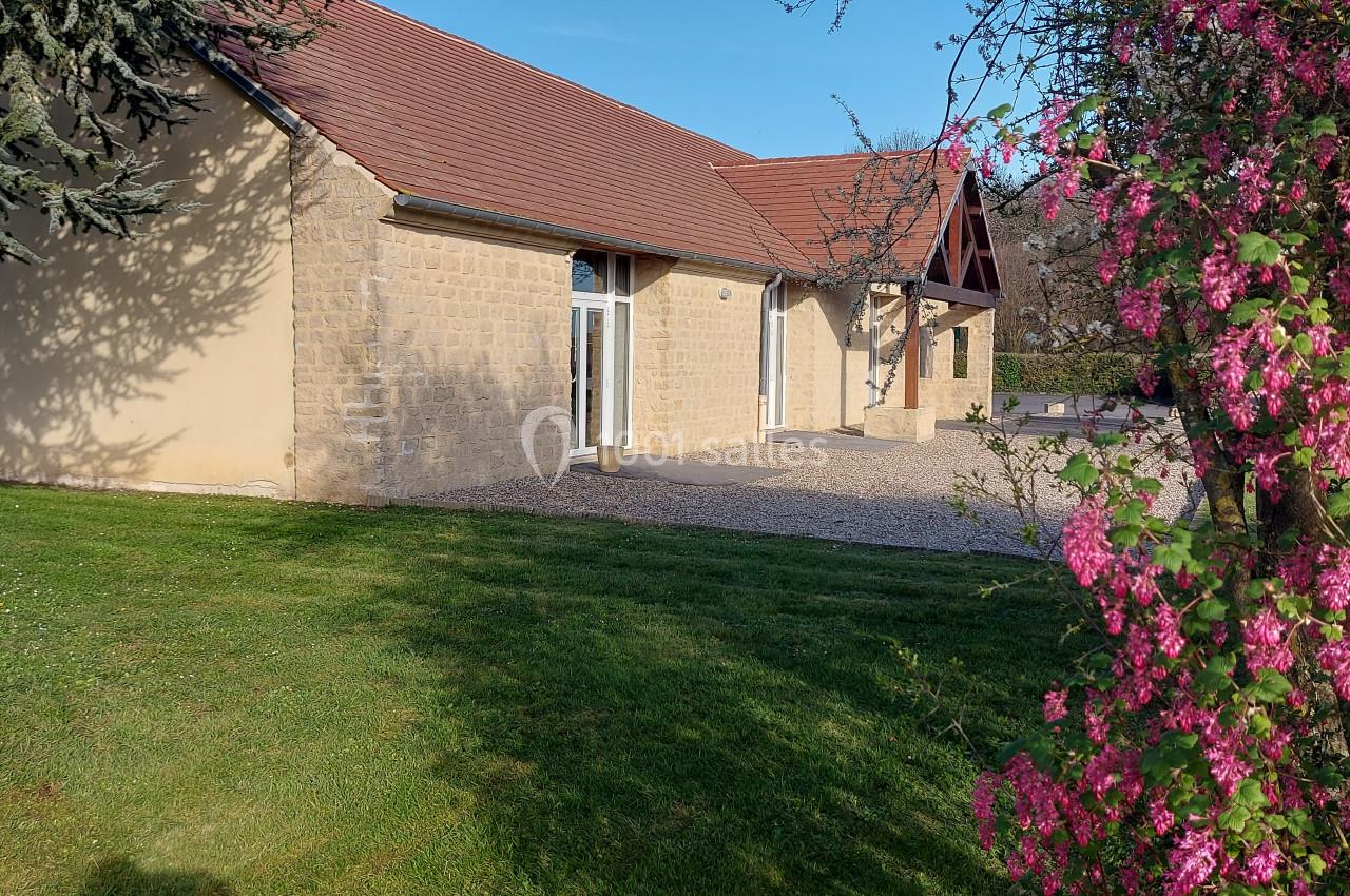 Bâtiment en pierre avec toit en tuiles, entouré de pelouse et d'arbustes fleuris sous un ciel dégagé.