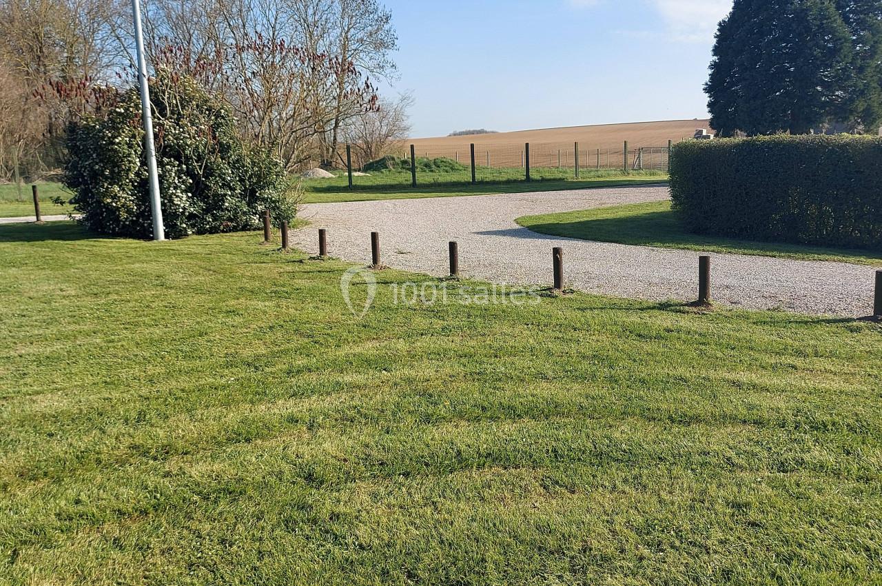 Pelouse verte bordée de poteaux en bois, chemin gravillonné et paysage rural avec arbres et champs en arrière-plan.