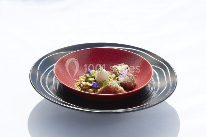 Assiette rouge sur plat noir contenant un plat raffiné avec fruits, fleurs comestibles et garnitures.