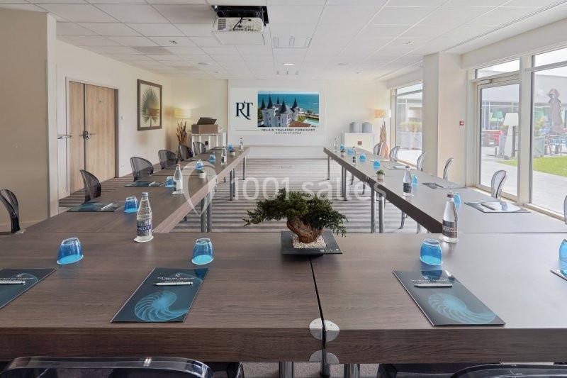 Salle de réunion lumineuse avec tables disposées en U, chaises transparentes et vue sur l'extérieur par de grandes baies…