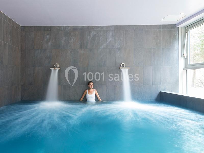 Femme se relaxant dans une piscine intérieure avec jets d'eau, entourée de murs en carrelage gris.