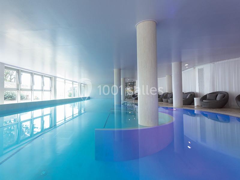 Piscine intérieure lumineuse avec colonnes blanches, grandes baies vitrées et fauteuils en osier sur le côté.