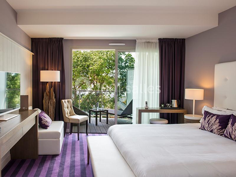 Chambre moderne avec lit double, bureau, fauteuils, et balcon donnant sur un espace extérieur verdoyant.