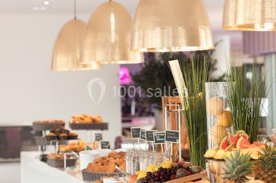 Salle de restaurant lumineuse avec tables dressées, chaises violettes, luminaires dorés et grandes baies vitrées.