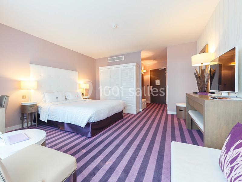 Chambre d'hôtel lumineuse avec lit double, mobilier moderne, moquette violette et éclairage chaleureux.
