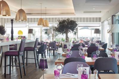 Salle de restaurant lumineuse avec tables dressées, chaises violettes, luminaires dorés et grandes baies vitrées.